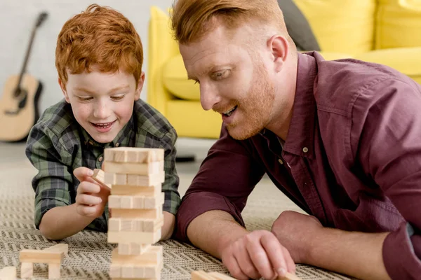 Feliz pelirroja padre e hijo acostado en la alfombra y jugando con bloques de madera - foto de stock
