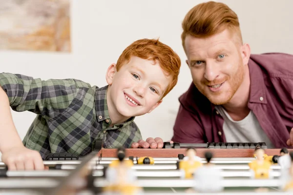 Feliz ruiva pai e filho sorrindo para a câmera enquanto joga futebol de mesa juntos em casa — Fotografia de Stock