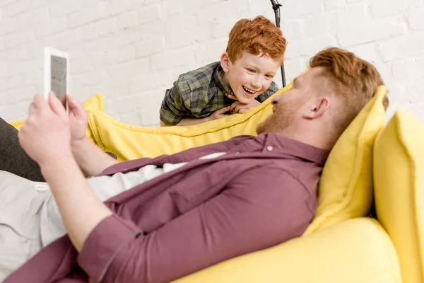 Glücklicher rothaariger Mann mit digitalem Tablet und Blick auf niedlichen kleinen Sohn — Stockfoto