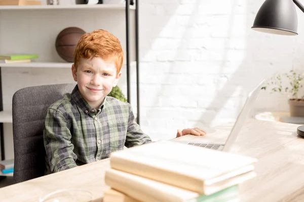 Netter kleiner rothaariger Junge lächelt in die Kamera, während er mit Büchern und Laptop am Tisch sitzt — Stockfoto