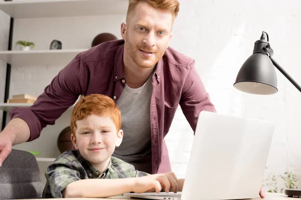 Sonriente padre e hijo usando el ordenador portátil juntos y mirando a la cámara en casa - foto de stock