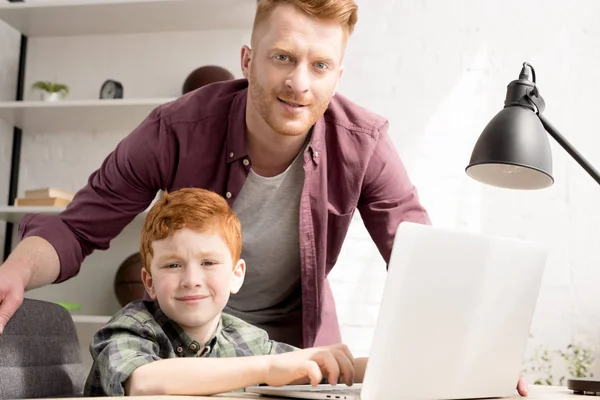 Sonriente padre e hijo usando el ordenador portátil juntos y mirando a la cámara en casa — Stock Photo