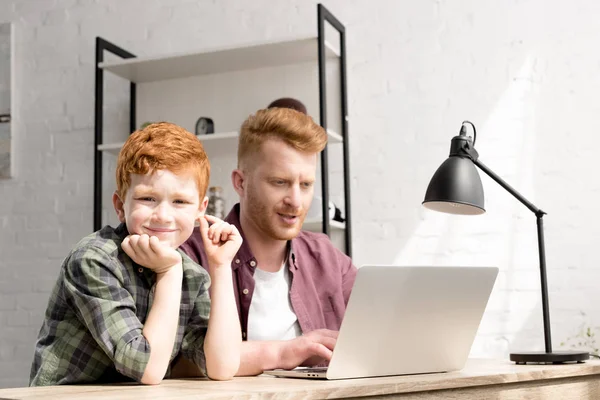 Милый мальчик улыбается в камеру во время использования ноутбука с отцом дома — стоковое фото