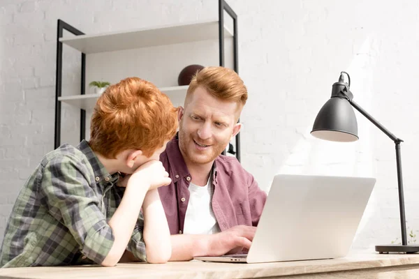 Padre e figlio felici che si sorridono mentre usano il computer portatile insieme a casa — Foto stock