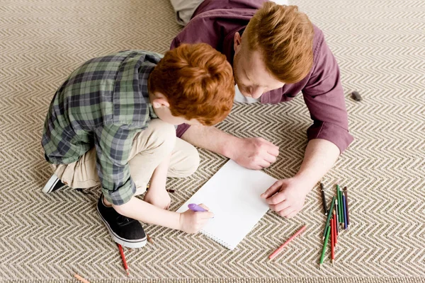 Vue aérienne du père roux et du fils dessinant ensemble sur le tapis — Photo de stock