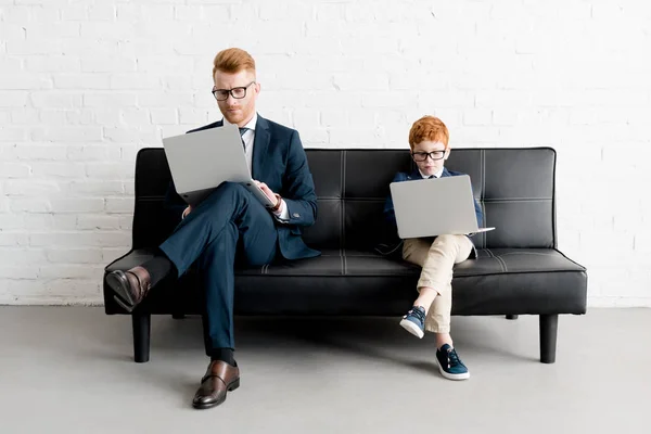 Padre serio e hijo pequeño hombres de negocios con anteojos y el uso de computadoras portátiles - foto de stock