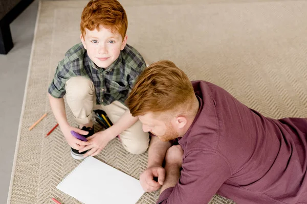 High angle view of cute little boy smiling at camera while drawing with father at home — Stock Photo