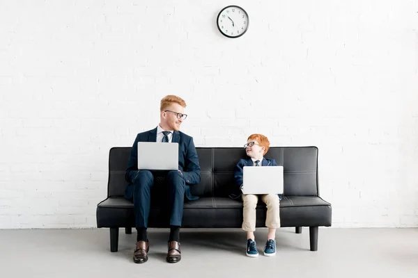 Lächelnde Vater-Sohn-Geschäftsleute in Brillen, die Laptops benutzen und einander anschauen — Stockfoto