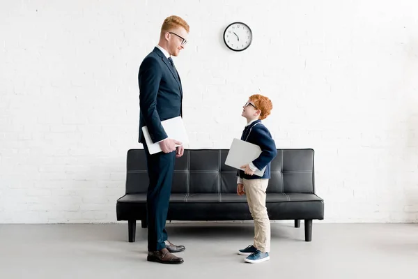 Vista lateral de adultos y preadolescentes hombres de negocios sosteniendo computadoras portátiles y sonriendo entre sí - foto de stock