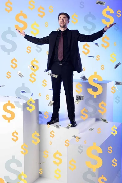 Homem de negócios sorridente de terno em pé no bloco branco com notas de dólar caindo e símbolos ao redor — Fotografia de Stock