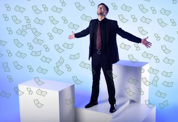 Uomo d'affari in completo nero con braccia tese in piedi su blocco bianco con banconote in dollaro in caduta su blu — Foto stock