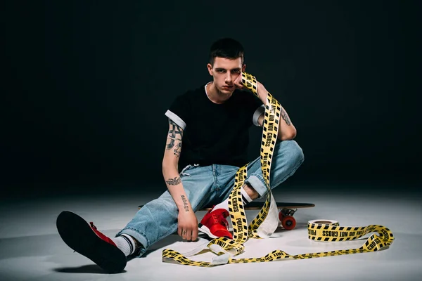 Hipster man sitting on longboard in yellow tape on dark background — Stock Photo