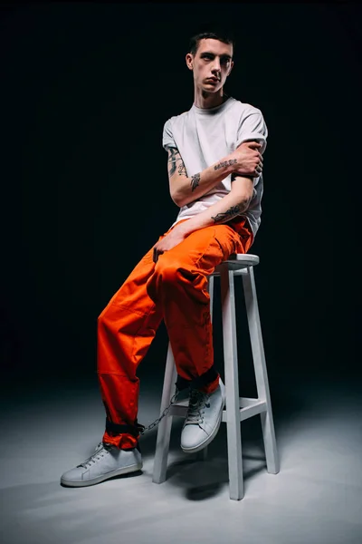 Homme tatoué portant un pantalon orange assis sur un tabouret sur fond sombre — Photo de stock