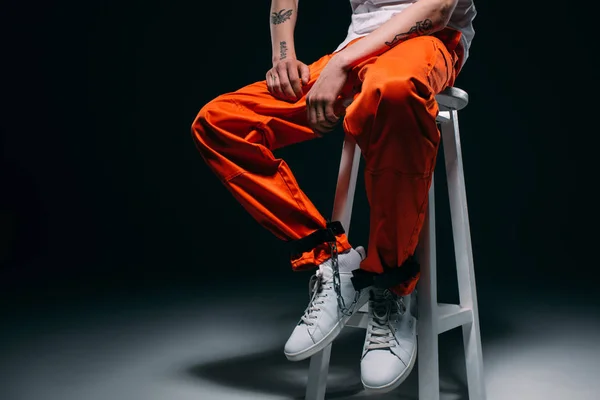 Cropped view of man in prison uniform with cuffs on legs sitting on stool on dark background — Stock Photo