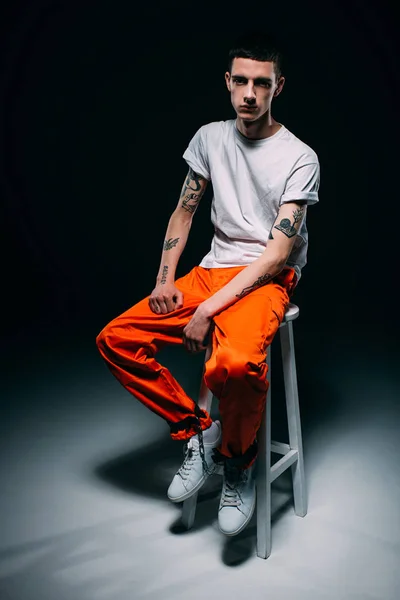 Serious man in orange uniform with cuffs on legs sitting on stool on dark background — Stock Photo