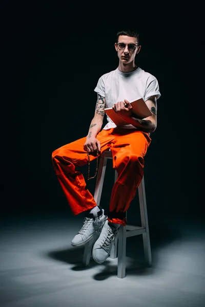 Uomo serio in uniforme arancione con polsini sulle gambe lettura bibbia e seduta su sgabello su sfondo scuro — Foto stock
