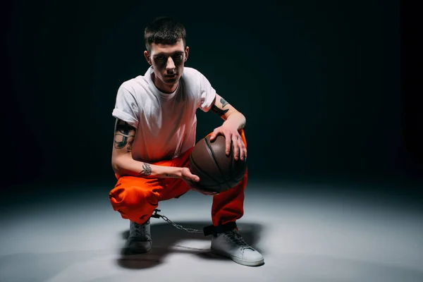 Hombre en pantalones y puños naranjas sosteniendo pelota de baloncesto sobre fondo oscuro - foto de stock