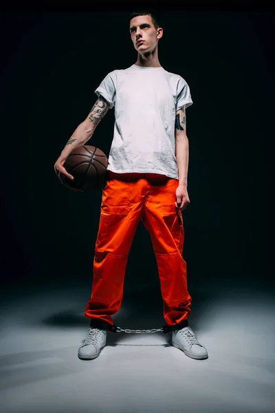 Mann in Gefängnisuniform und Handschellen hält Basketballball auf dunklem Hintergrund — Stockfoto