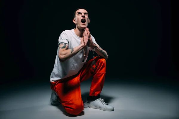 Screaming man in prison uniform and cuffs holding rosary and praying on dark background — Stock Photo