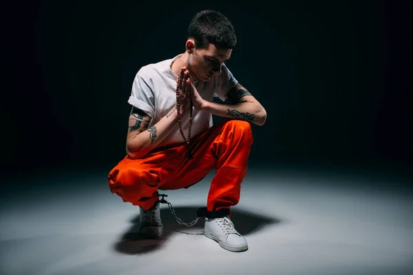 Sad male prisoner in orange pants and cuffs holding rosary and praying on dark background — Stock Photo