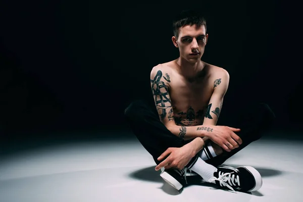 Молодой человек с татуированными руками сидит на полу на темном фоне — стоковое фото