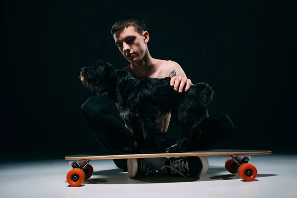 Cão preto em pé no longboard por seu proprietário no fundo escuro — Fotografia de Stock