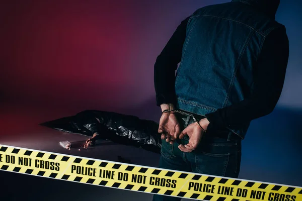 Criminal in handcuffs by body in trash bag behind police tape on dark background — Stock Photo