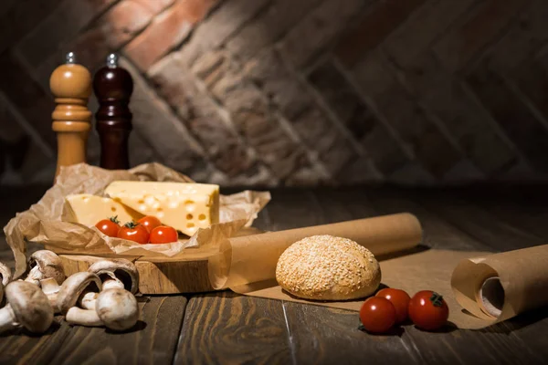 Close up view of fresh cherry tomatoes, cheese, mushrooms and loaf of bread on baking paper on wooden tabletop — Stock Photo