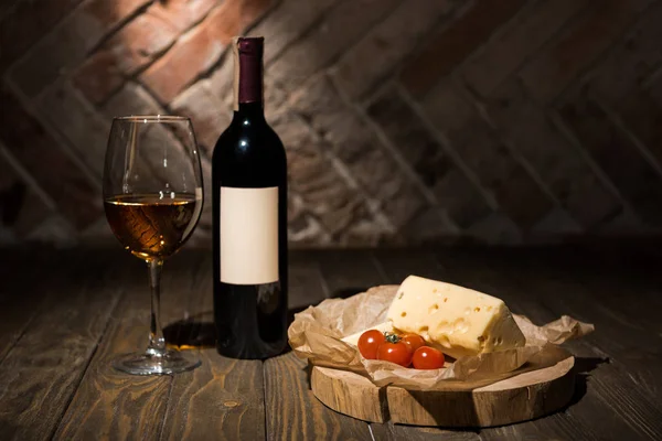 Primer plano vista de botella y copa de vino con queso y tomates cherry sobre papel de hornear en tocón decorativo de madera - foto de stock