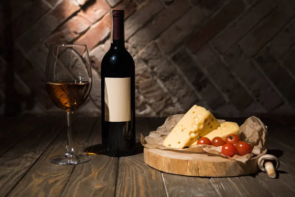 Primer plano vista de botella y copa de vino con queso y tomates alegres sobre papel de hornear en tocón decorativo de madera - foto de stock
