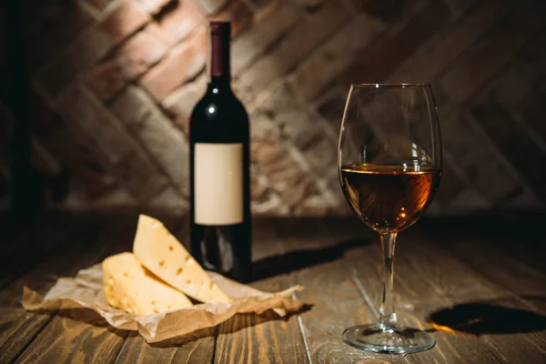 Vista ravvicinata di bottiglia e bicchiere di vino con formaggio su carta da forno su tavolo in legno — Foto stock