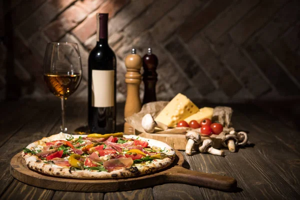 Foco seletivo de pizza italiana, especiarias, queijo, tomate cereja e garrafa de vinho na superfície de madeira — Fotografia de Stock