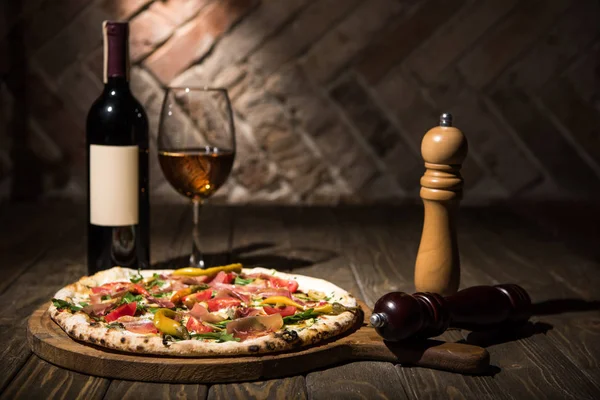 Enfoque selectivo de la pizza italiana, especias en molinillos, botella y copa de vino en la mesa de madera - foto de stock