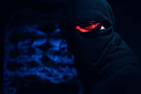 Obscured view of hacker in eyeglasses and mask looking at camera with cables on background — Stock Photo