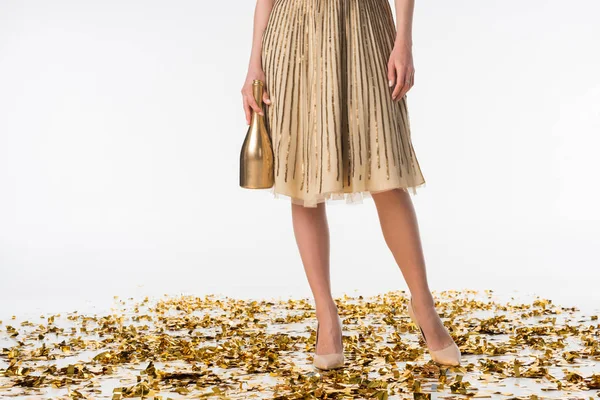Image recadrée de fille debout sur confettis en jupe et tenant bouteille de champagne — Photo de stock