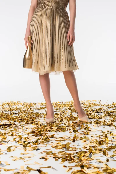 Imagen recortada de la mujer de pie sobre confeti en vestido y la celebración de la botella de oro de champán - foto de stock