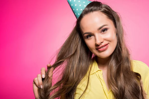 Atractiva mujer con sombrero de fiesta tocando pelo aislado en rosa - foto de stock