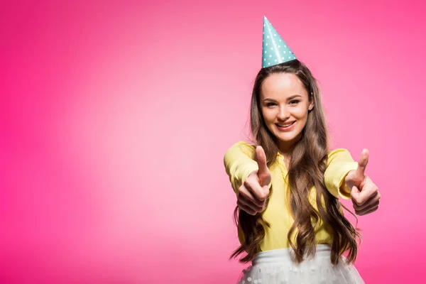 Atractiva mujer con sombrero de fiesta que muestra los pulgares hacia arriba aislado en rosa - foto de stock