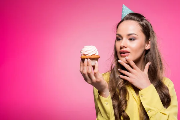 Attraktive Frau mit Party-Hut hält Cupcake isoliert auf rosa — Stockfoto