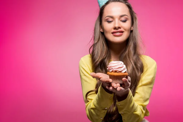 Lächeln attraktive Frau mit Party-Hut hält Cupcake isoliert auf rosa — Stockfoto