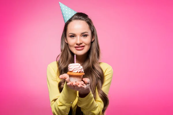 Attraktive Frau mit Partyhut, Cupcake in der Hand und Blick in die Kamera isoliert auf rosa — Stockfoto