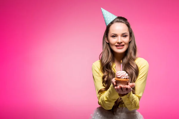 Attraktive Frau mit Party-Hut hält Cupcake mit Kerze isoliert auf rosa — Stockfoto