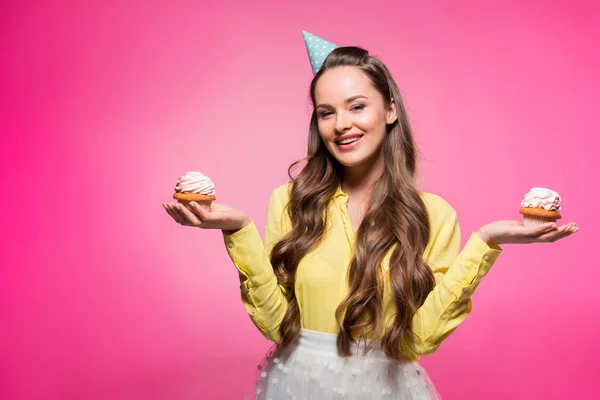 Attraktive Frau mit Partyhut hält leckere Cupcakes isoliert auf rosa — Stockfoto