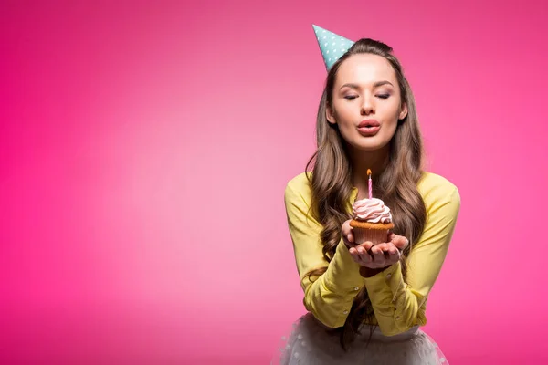 Attractive woman with party hat holding cupcake and blowing out candle isolated on pink — Stock Photo