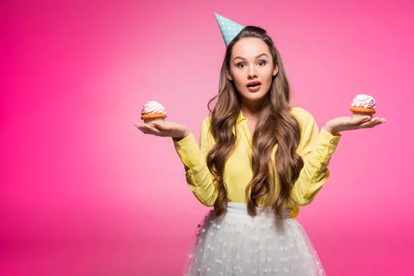 Attractive woman with party hat holding cupcakes isolated on pink — Stock Photo