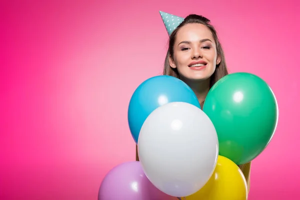 Attractive woman with party hat looking out from balloons isolated on pink — Stock Photo