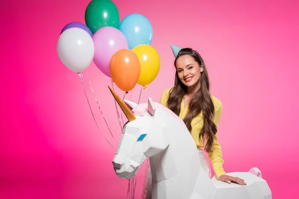 Atractiva mujer con sombrero de fiesta, globos y unicornio juguete aislado en rosa - foto de stock