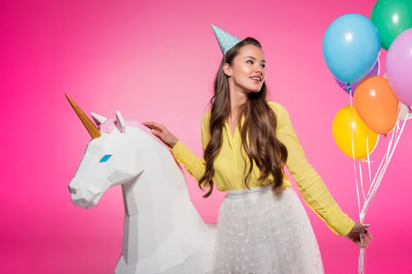 Hermosa chica con sombrero de fiesta, globos y unicornio juguete aislado en rosa - foto de stock
