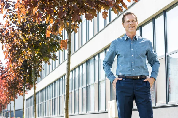 Happy mature businessman posing with hands in pockets near office building — Stock Photo