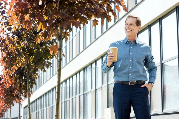 Sonriente hombre de negocios de pie con café para ir cerca del edificio de oficinas - foto de stock